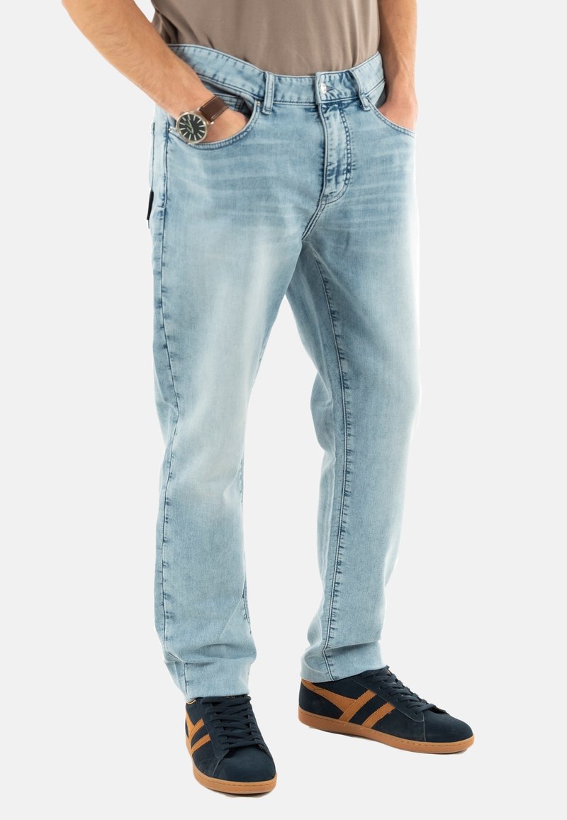 Jeans de mezclilla azul claro con un corte relajado, que cuentan con cinco bolsillos y una textura desgastada. Combinados con zapatillas negras y detalles en marrón.