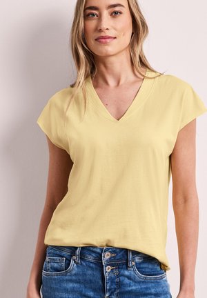 Nainen pukeutuneena vaaleankeltaiseen V-kaula-aukkoinen lyhythihainen yläosa ja sinisiin farkkuihin, seisoo yksivärisen vaalean taustan edessä.