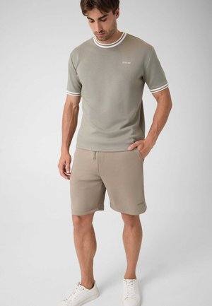 Homme portant un short beige et un T-shirt kaki assorti avec des poignets rayés blancs et des baskets blanches, debout avec une main dans la poche.