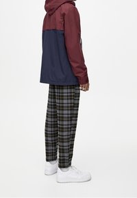 Bordeauxrode en marineblauwe gewatteerde jas met capuchon en een losse pasvorm, gecombineerd met grijs-gele geruite broeken en witte sneakers, die casual streetwear benadrukken.