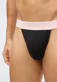 Close-up van een persoon in zwarte onderbroeken met een lichtroze Calvin Klein tailleband. Zichtbare huid, hand en bovenbenen.