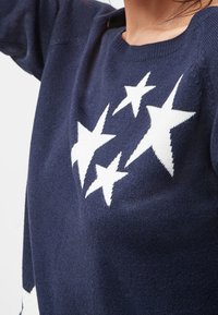 Pull en maille bleu marine avec des motifs d'étoiles blanches sur la poitrine, porté par une personne levant le bras, le cou et une partie du visage visibles.