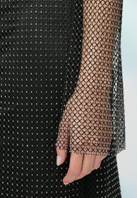 Fekete hálós ruha ujj, gyémánt mintával, fémes részletekkel. Közeli kép a kézről, sima textúrával és természetes körmökkel.