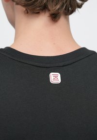 Μαύρο βαμβακερό t-shirt με στρογγυλή λαιμόκοψη· κόκκινη και λευκή τετράγωνη ετικέτα ραμμένη στο πίσω μέρος με στυλιζαρισμένα κόκκινα γράμματα.