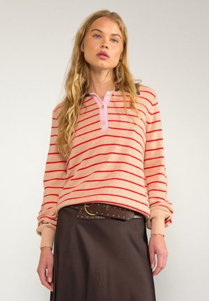 Jeune femme aux longs cheveux blonds portant un pull rayé rose et rouge et une jupe marron foncé avec une ceinture cloutée.