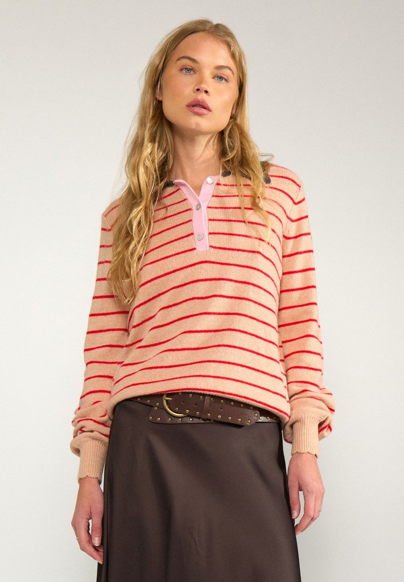 Jeune femme aux longs cheveux blonds portant un pull rayé rose et rouge et une jupe marron foncé avec une ceinture cloutée.