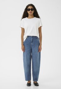 T-shirt bianco a maniche corte abbinato a jeans in denim blu a vita alta e larghi e scarpe appiattite nere. Il modello indossa occhiali da sole neri.