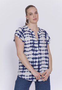 Kurze Ärmel Bluse in Weiß mit einem blauen Batikmuster. Sie hat einen V-Ausschnitt und Rüscheärmel. Leichter Stoff mit lockerer Passform.