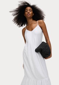 Weißes ärmelloses Kleid mit plissierten Details, ausgestelltem Saum und dünnen Trägern. Das Model hält eine strukturierte schwarze geflochtene Clutch. Neutraler Hintergrund.