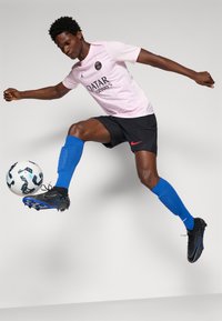 Futbolo žaidėjas šviesiai rožinėje maikutėje, juodose šortuose, mėlynose kojinėse ir juoduose sportbačiuose, atlieka smūgį baltu ir juodu futbolo kamuoliu.