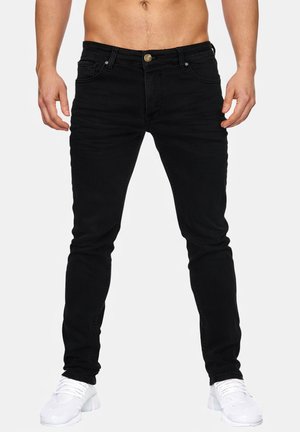 Homme portant un jean noir slim et des chaussures de sport blanches, debout les mains relâchées le long du corps sur un fond uni.