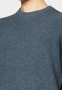 Gros plan sur une personne portant un pull bleu en tricot côtes avec des détails de texture et de couture.