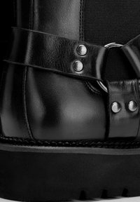 Bota de cuero negro con diseño brogue, secciones de tela texturizada, detalles metálicos y una suela negra gruesa.