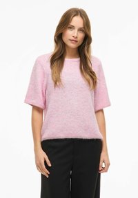 Rozā, īsroku džemperis no mīksta, pūkaina materiāla ar brīvu piegriezumu, apaļu izgriezumu un smalkām tekstūras variācijām.