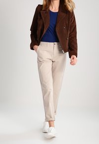 Kvinna som går klädd i en brun jacka, marinblå skjorta, beige uppvikta byxor och vita sneakers mot en enfärgad ljus bakgrund.