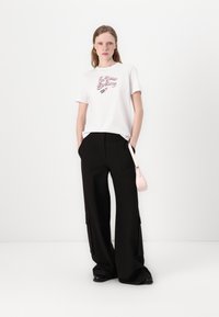 Tricou alb cu text grafic roz "În Visurile Tale", purtat cu pantaloni cargo negri cu talie înaltă și o geantă mică roz tip crossbody.