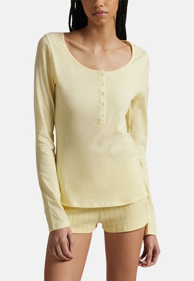 Femme portant un haut à manches longues boutonné jaune pâle et un short texturé assorti sur un fond clair uni.