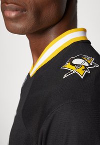 Fekete hoki mez textúrált felülettel, sárga és fehér csíkos gallérral, valamint díszes pingvin logóval a vállon.