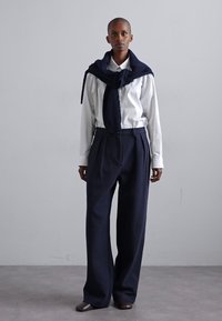Modella che indossa una camicia bianca con bottoni e pantaloni a vita alta di colore blu navy, larghi. Un maglione in maglia blu navy è posato sulle spalle.