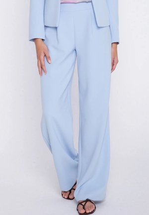 Jasnoniebieskie, szerokie spodnie z wysokim stanem noszone z dopasowaną marynarką i czarnymi sandałami na cienkich paskach na stojącej osobie.