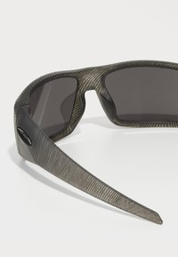 Sonnenbrille mit einem gestreiften Rahmen in Grau und Schwarz, dunklen Gläsern und einem sportlichen, aerodynamischen Design. Schwarze Hardware-Details an den Bügeln.
