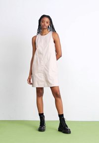 Vestido sin mangas beige con corte recto, que presenta una costura central y un diseño minimalista. Combinado con botas de combate negras y calcetas.