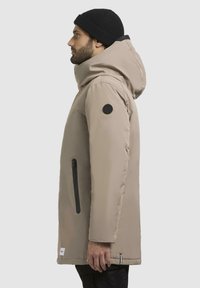 Beige parka med hætte, sorte detaljer, sidelomme med lynlås og logo-patch på armen. Glat tekstur og lige snit.