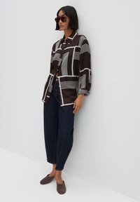 Camicia a maniche lunghe con bottoni di colore marrone scuro e motivi geometrici bianchi, abbinata a jeans a gamba larga blu scuro e scarpe slip-on marroni.