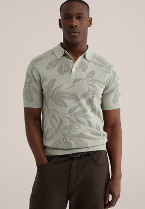 Vyras dėvi šviesiai žalią trumpomis rankovėmis megztą polo marškinėlį su lapų raštu ir tamsiai rudos spalvos kelnes, stovi viena ranka kišenėje.