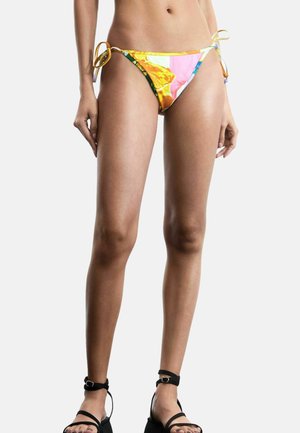 Naine kannab värvilisi külgedest lipsuga bikiinipõhjust ja musti platvorm-sandaale pahkluu rihmaga, seisab ühevärvilise valge tausta ees.