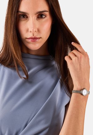 Montre argentée avec un cadran rond blanc et un bracelet en maille texturée. La personne qui la porte a de longs cheveux bruns, se détachant sur un fond en tissu bleu doux.