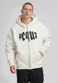 Witte zip-up hoodie met zwarte "pequs" tekst, voorzakken, geribbelde boorden en een gevoerde capuchon. Zacht stof en relaxte pasvorm.