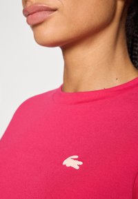 Tett rosa bomull t-skjorte med rund hals og korte ermer, med et lite lysebeige brodert logo på brystet.