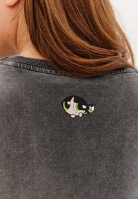 Světle šedý svetr s kulatou, barevnou nášivkou kreslené postavičky s zelenýma očima a černo-bílými detaily na zadní straně.