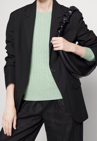 Zwarte blazer met een enkele knoop, gedragen over een lichtgroene kabelgebreide trui. Zwarte handtas met een gestructureerde band. Donkere, losvallende broek.