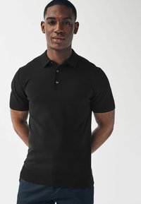 Černé polo tričko z pletené tkaniny. Obsahuje dvouknoflíkovou légu, krátké rukávy a žebrovaný lem. Model stojí se zkříženýma rukama za zády.