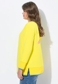 Haut à manches longues jaune avec un ourlet arrondi, accents bleus aux poignets et un détail à motif rose dans le dos. Porté avec un jean en denim.