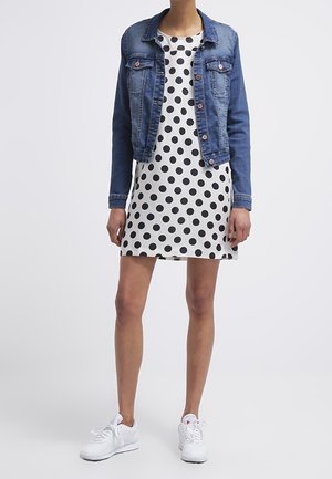 Femme portant une robe blanche à pois noirs, une veste en jean bleue et des baskets blanches, debout devant un fond uni.