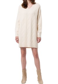 Cremfarbener gestrickter Pulloverkleid mit V-Ausschnitt, ausgestattet mit Rautenmuster und gerippten Bündchen. Kombiniert mit beigen Stiefeletten.