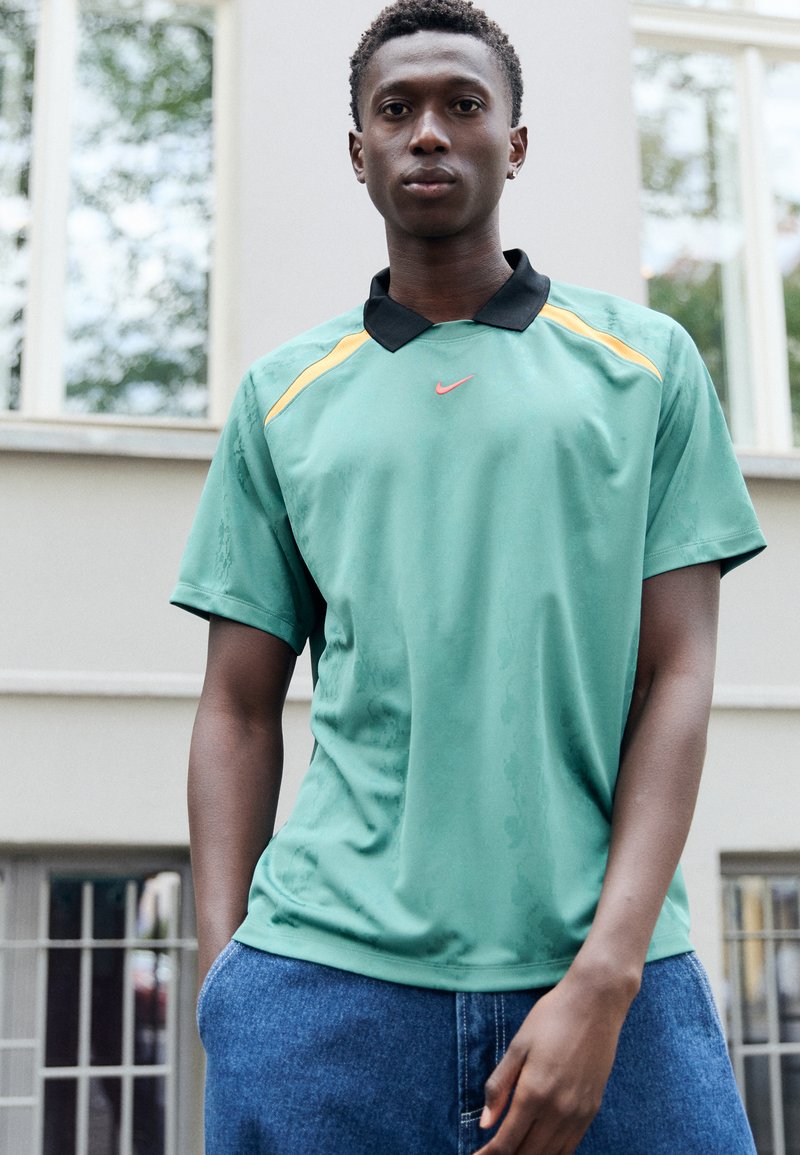 Polo shirt Nike vert avec un col noir et des accents jaunes. Le tissu semble léger et a une texture lisse. Le logo est en rouge.
