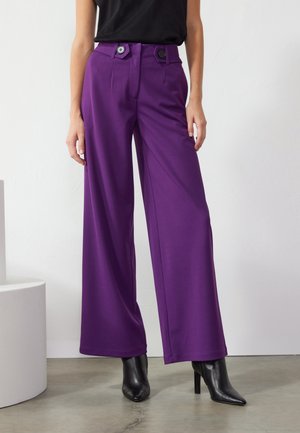Femme portant un pantalon violet taille haute à jambes larges avec de gros boutons noirs, un haut noir et des bottes noires à talons pointus, debout à l'intérieur.