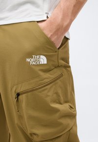 Το χέρι ενός ατόμου στην τσέπη παντελονιών North Face ελιάς με μπροστινή τσέπη με φερμουάρ και μερικά ορατό λευκό τοπ σε λευκό φόντο.