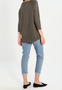 Übergroßes olivgrünes Oberteil mit Dreiviertelärmeln, verzierten Schultern, kombiniert mit hellblauen, hochgekrempelten Jeans und schwarzen Ballerinas.