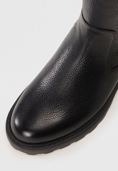 Bottine en cuir noir avec une finition texturée, présentant un embout arrondi et une semelle en caoutchouc flexible. Couture visible sur les côtés.