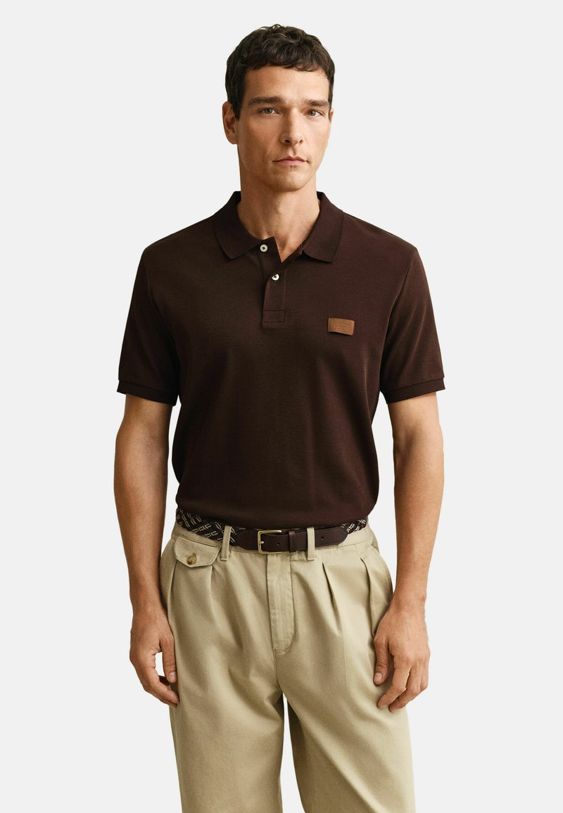 Man draagt een donkerbruin kortärmelig poloshirt, ingestopt in beige geplooide broek met een riem met patroon, tegen een effen witte achtergrond.