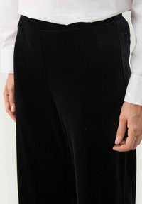 Pantaloni negri din catifea cu o textură netedă, având un design cu picior drept și o talie elastică. Vedere îndeaproape a materialului și a potrivirii.