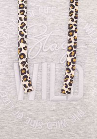 Sudadera gris con "Stay WILD" bordado en una textura suave. Presenta cordones con estampado de leopardo y un patrón circular de frases alrededor del cuello.