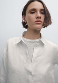 Wit linnen overhemd met knopen en een zachte kraag, gedragen over een licht geribd shirt, gecombineerd met een zilveren ketting en oorbellen in hoepelvorm.