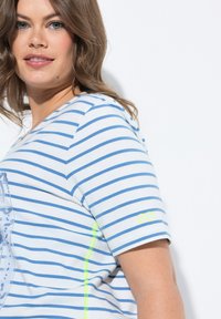 Gestreiftes T-Shirt aus weißem und blauen Stoff mit kurzen Ärmeln. Mit einem grünen Akzentnähte und Logo am Ärmel. Weicher Baumwollstoff.