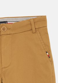 Tan cotton trousers with a button closure, side pocket, and subtle logo stripe detailing in red, white, and blue on the pocket.